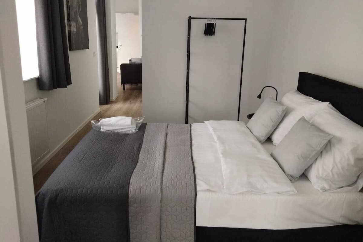 Un lit bien fait, avec du linge de lit blanc et une couverture grise, une tête de lit noire et une table de chevet avec une lampe.