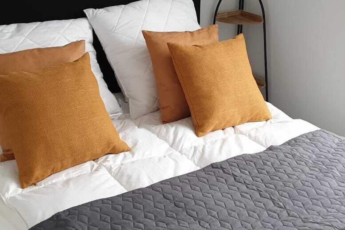Un lit bien fait, avec une tête de lit noire, des oreillers blancs et moutarde, et une couvre-lit grise matelassée.