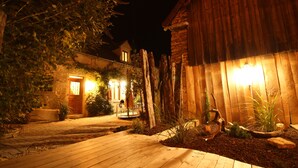 Eine Holzterrasse führt zu einem Steinhaus mit Tür und Fenstern, umgeben von Pflanzen und einem Holzzaun.