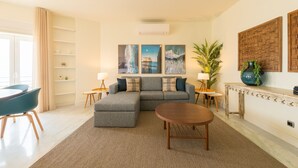 Een moderne woonkamer met een hoekbank, een ronde salontafel en een eethoek met stoelen. Er zijn wandplanken en een grote potplant.