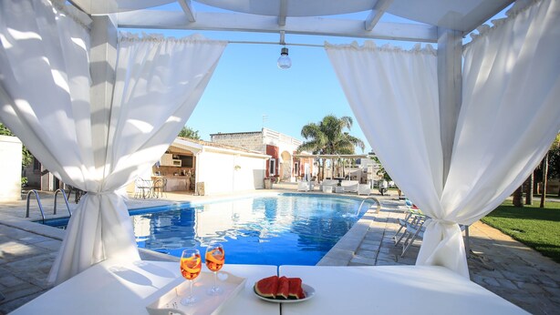 A poolside table with a watermelon and two glasses of orange juice, a white canopy, and a clear view of a swimming pool and buildings in the background.