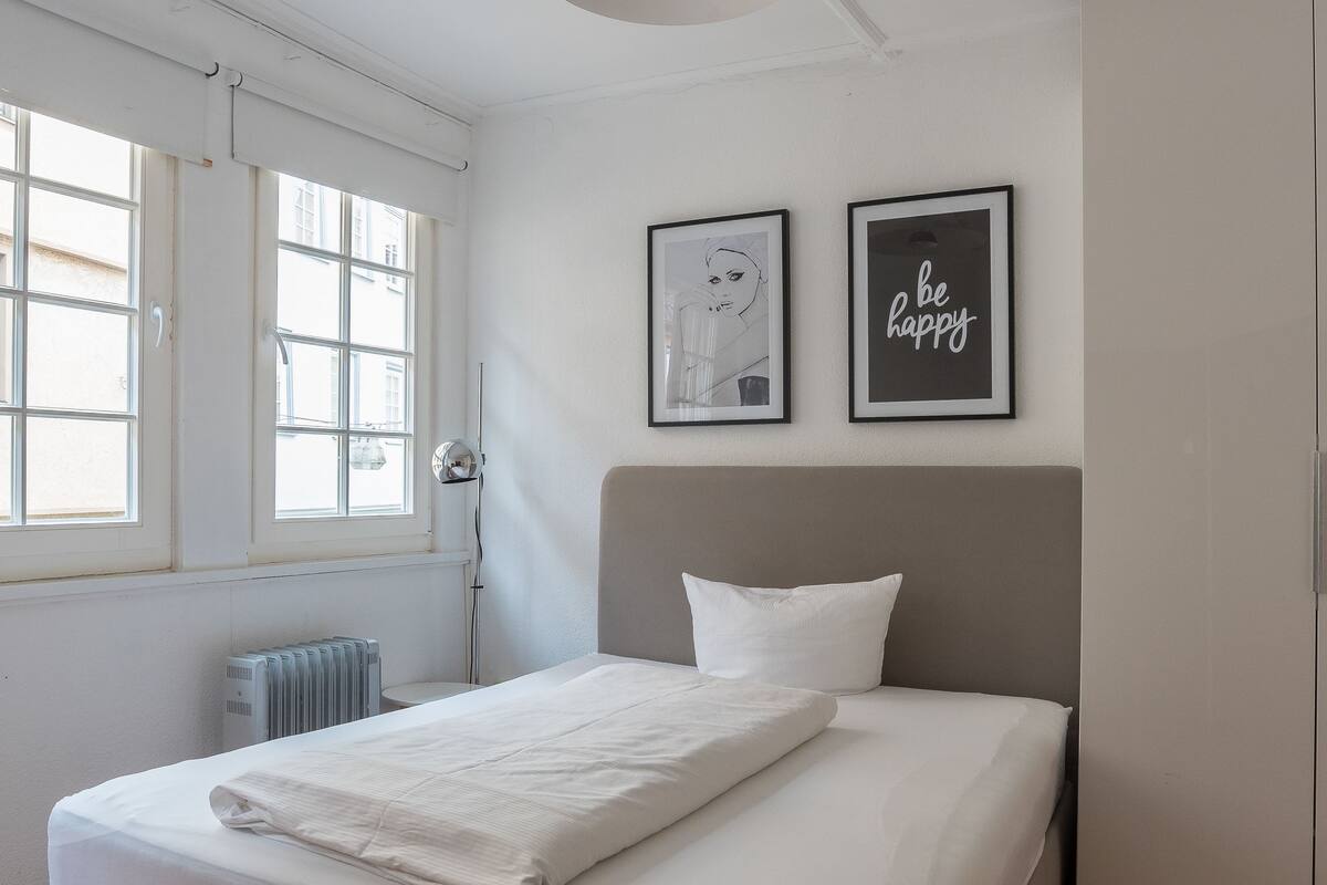 Quarto com cama, radiador, janela e duas molduras na parede.