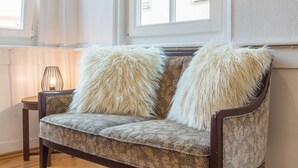 A sofa with fluffy pillows, a small table with a lamp, and a window with a view of buildings. - Privatzimmer in bester Altstadtlage (Tuebingen)