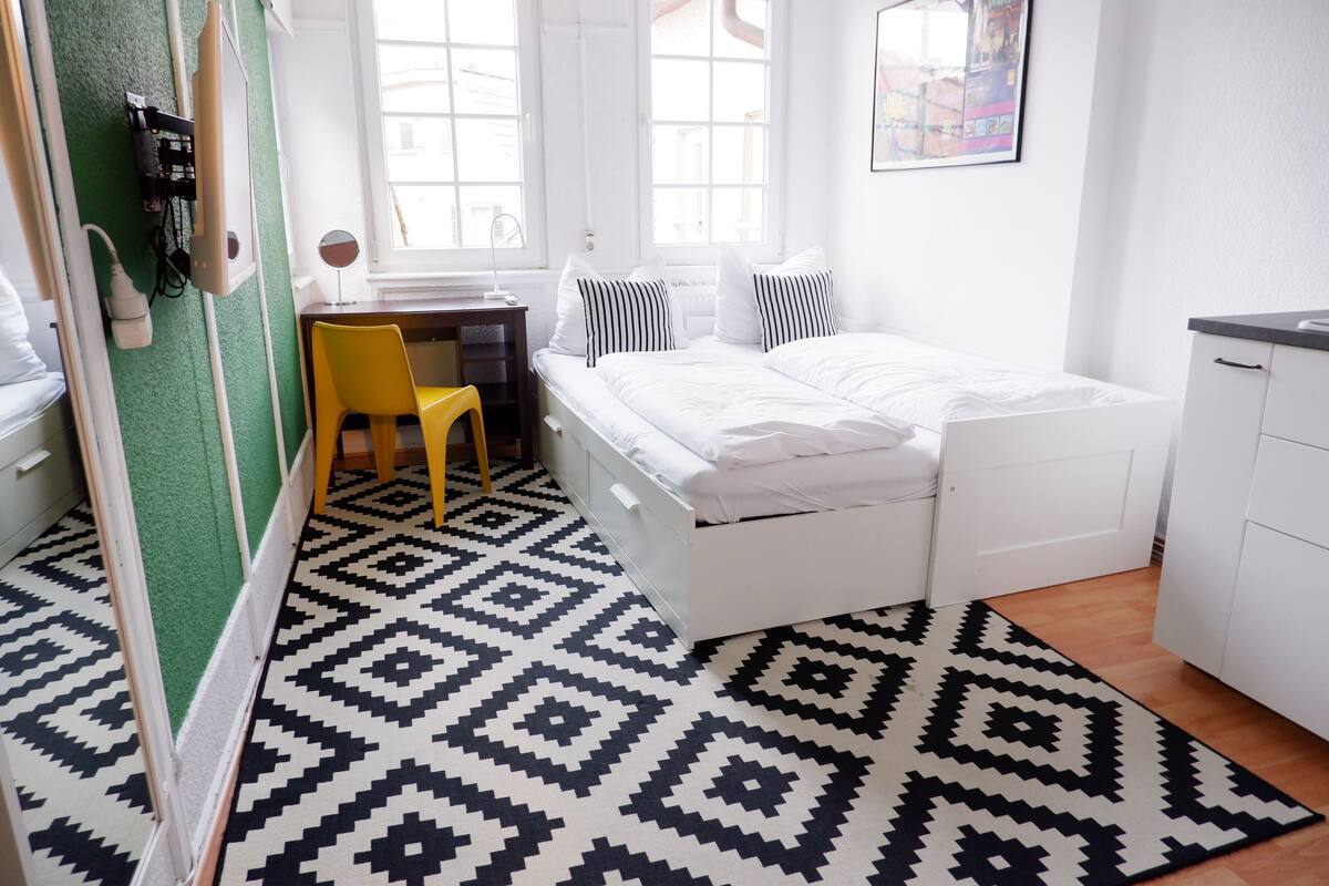 Quarto pequeno e luminoso com uma cama, uma escrivaninha e uma cadeira. Há janelas, um radiador e uma televisão presa à parede.