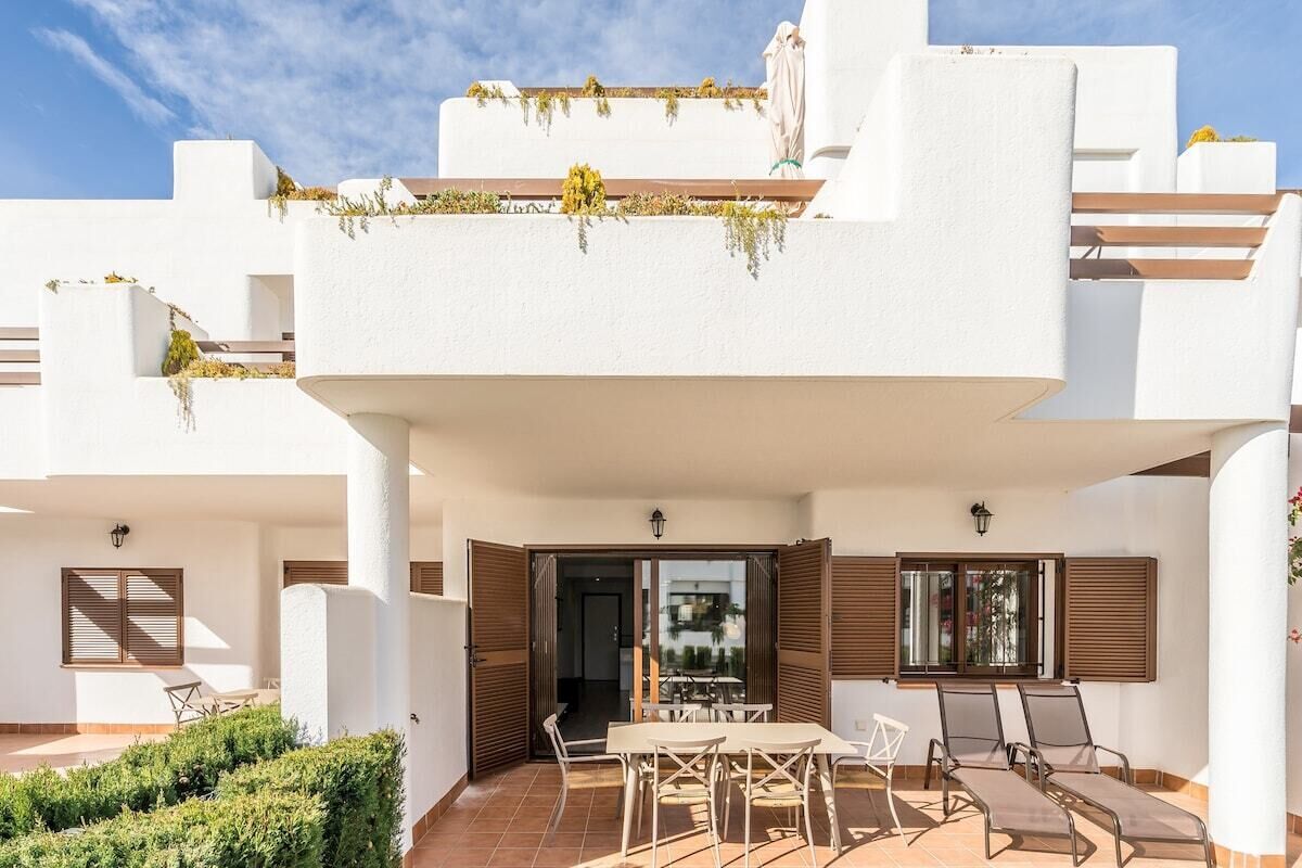 Un pati exterior modern amb taula i cadires, un jardí petit i un edifici amb balcons de color blanc.