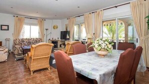 Ein Essbereich mit einem für vier Personen gedeckten Tisch, umgeben von Korbstühlen. Links befindet sich ein Fernseher auf einem Schrank, und rechts ein großes Fenster mit Vorhängen.