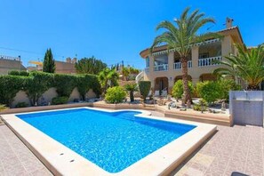 A swimming pool with a tiled deck, surrounded by a landscaped garden with palm trees and shrubs, and a two-story building with balconies. - CH Villa Jupiter La Marquesa Golf (Ciudad Quesada) (La Marquesa)