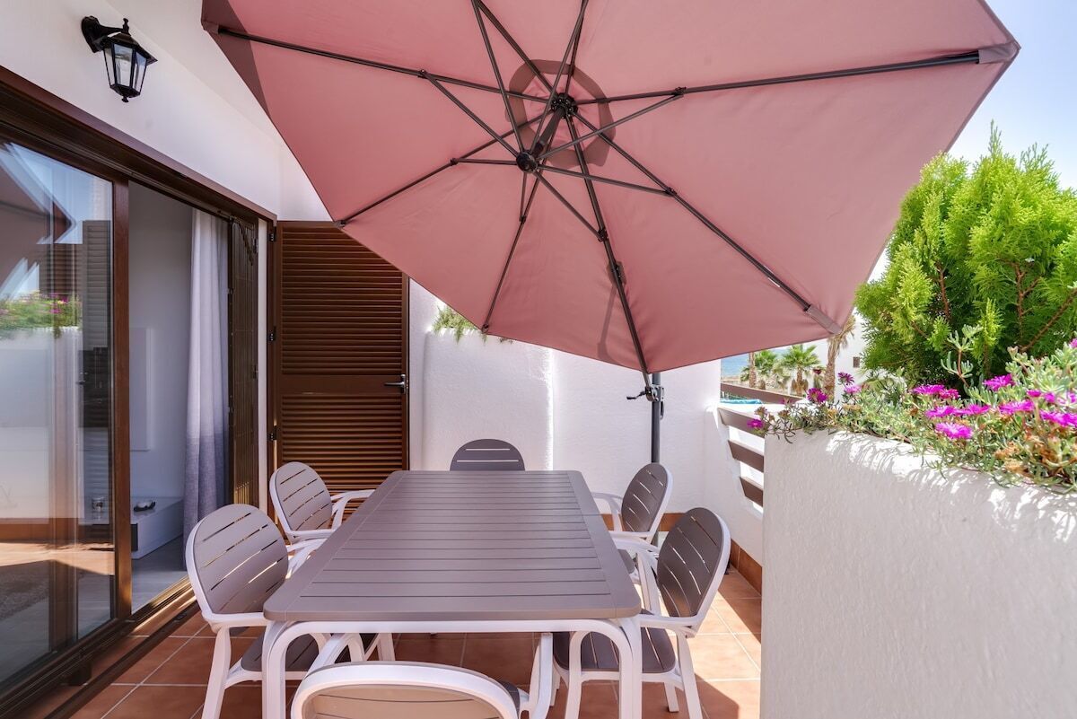 Un patio con mesa y sillas, un paraguas grande y vistas a plantas y flores.