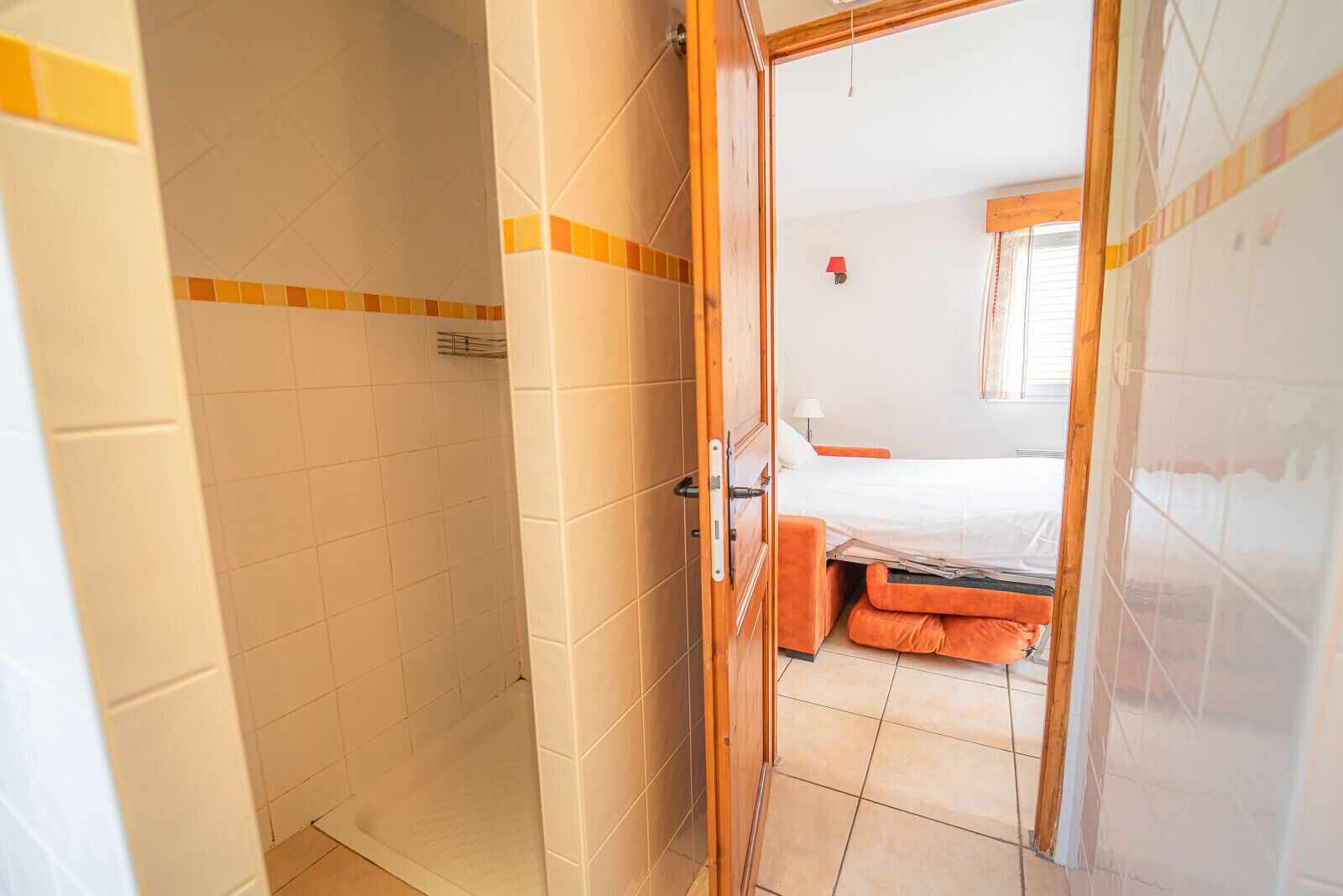A room with a shower, a bed with orange bedding, and a window with curtains.