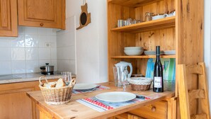 Une cuisine en bois avec une table à manger dressée pour deux personnes, agrémentée d’un panier de pain, d’une bouteille de vin et de divers ustensiles de cuisine.