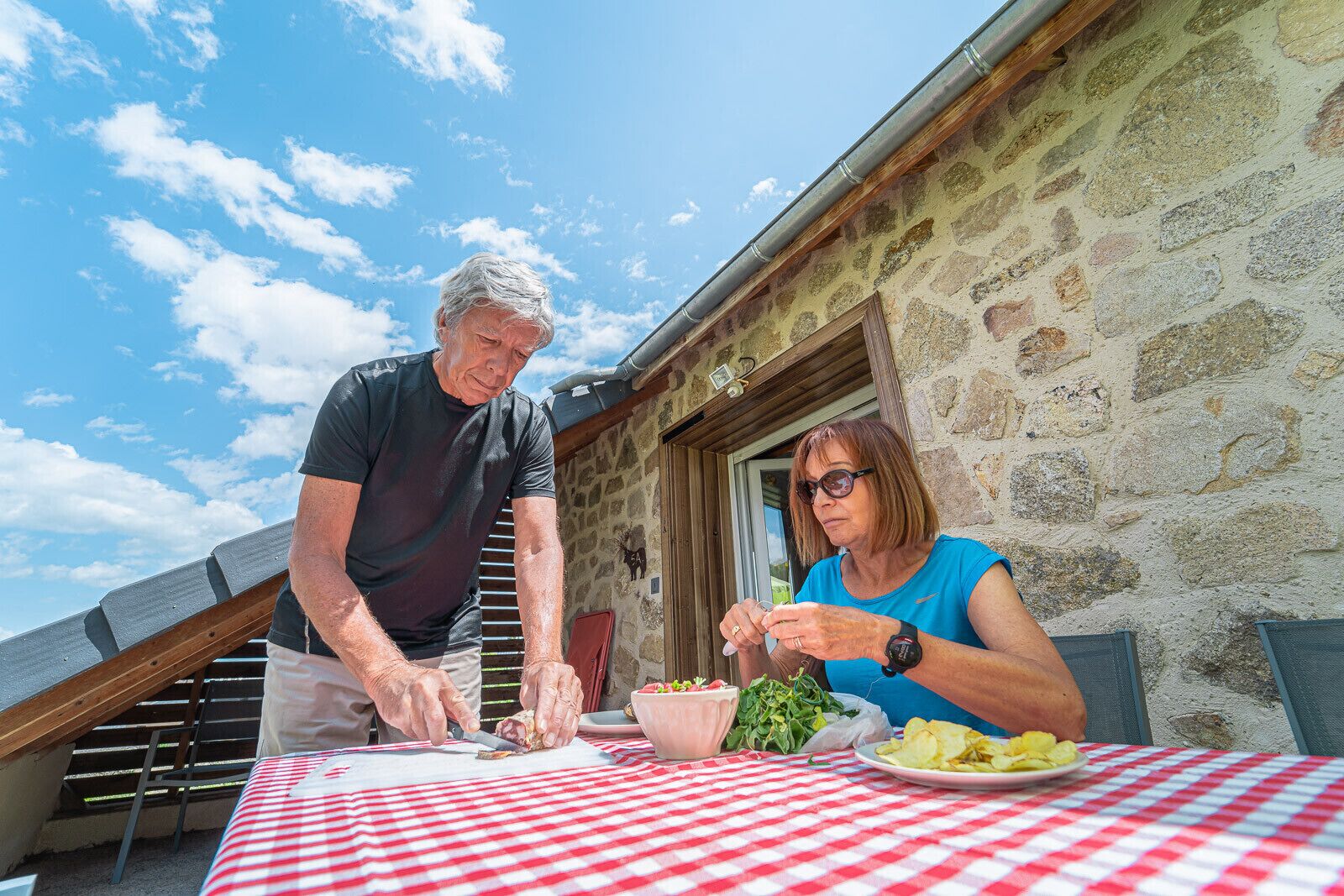 Two people dining at a table with a red and white checkered tablecloth, outdoors with a stone building in the background.