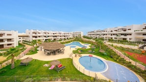 Un complex turístic amb diversos edificis blancs, una piscina central i una zona de jardí paisatjística amb palmeres i gandules.