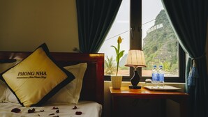 Une chambre d’hôtel avec un lit, un oreiller orné du mot « PHONG NHA », une table de chevet avec une lampe, deux bouteilles d’eau et une fenêtre donnant sur une montagne.