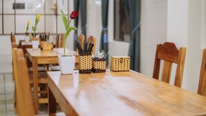 Une table de salle à manger en bois, sur laquelle est posé un vase blanc contenant une seule rose rouge, le tout entouré de récipients tressés contenant des ustensiles.