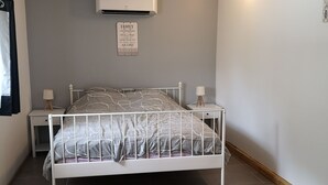 A bedroom with a white metal bed frame, grey patterned bedding, two bedside tables with lamps, a wall-mounted air conditioner, and a motivational quote sign.