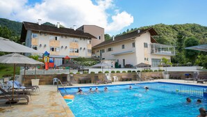 Ein Poolbereich mit Liegestühlen und Sonnenschirmen, umgeben von Gebäuden mit Balkonen und Blick auf eine hügelige Landschaft.