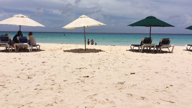 A beach with white sand and clear blue water. There are several beach umbrellas and lounge chairs.