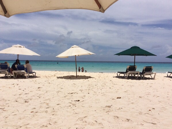 A beach with white sand and clear blue water. There are several beach umbrellas and lounge chairs.