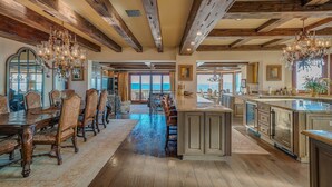 A spacious dining area with a long wooden table and ornate chairs, a central island with a bar, and a view of the sea through large windows. - Beach Front Executive Home (Long Beach)