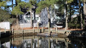 Un muelle de madera se adentra en una masa de agua tranquila, rodeado de altos pinos y una hilera de casas blancas con contraventanas verdes.