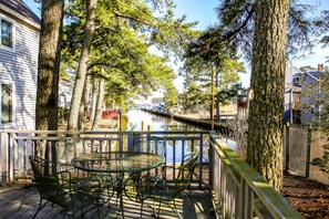 A wooden deck with a round table and chairs, surrounded by tall trees and overlooking a calm water body. - Blarney Shores -Waterfront -Pet Friendly - Balconies , No Linens Provided (Chincoteague)