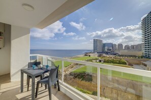 A balcony with a table and chairs, overlooking a coastal cityscape with buildings and a view of the sea.