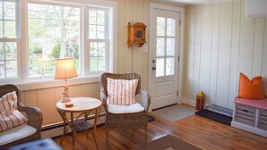 A cozy room with a wooden floor, two wicker chairs with striped cushions, a small round table with a lamp, a door with a mailbox, and a bench with orange cushions. - Three bedroom home- quick stroll to Nantucket Sound Beaches (Dennis Port)