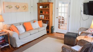 A living room with a grey sofa, orange cushions, a wooden bookshelf, wicker chairs, and a television mounted on the wall. - Three bedroom home- quick stroll to Nantucket Sound Beaches (Dennis Port)