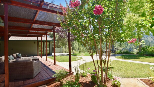 A covered outdoor seating area with a pergola, a lawn, and flowering plants.