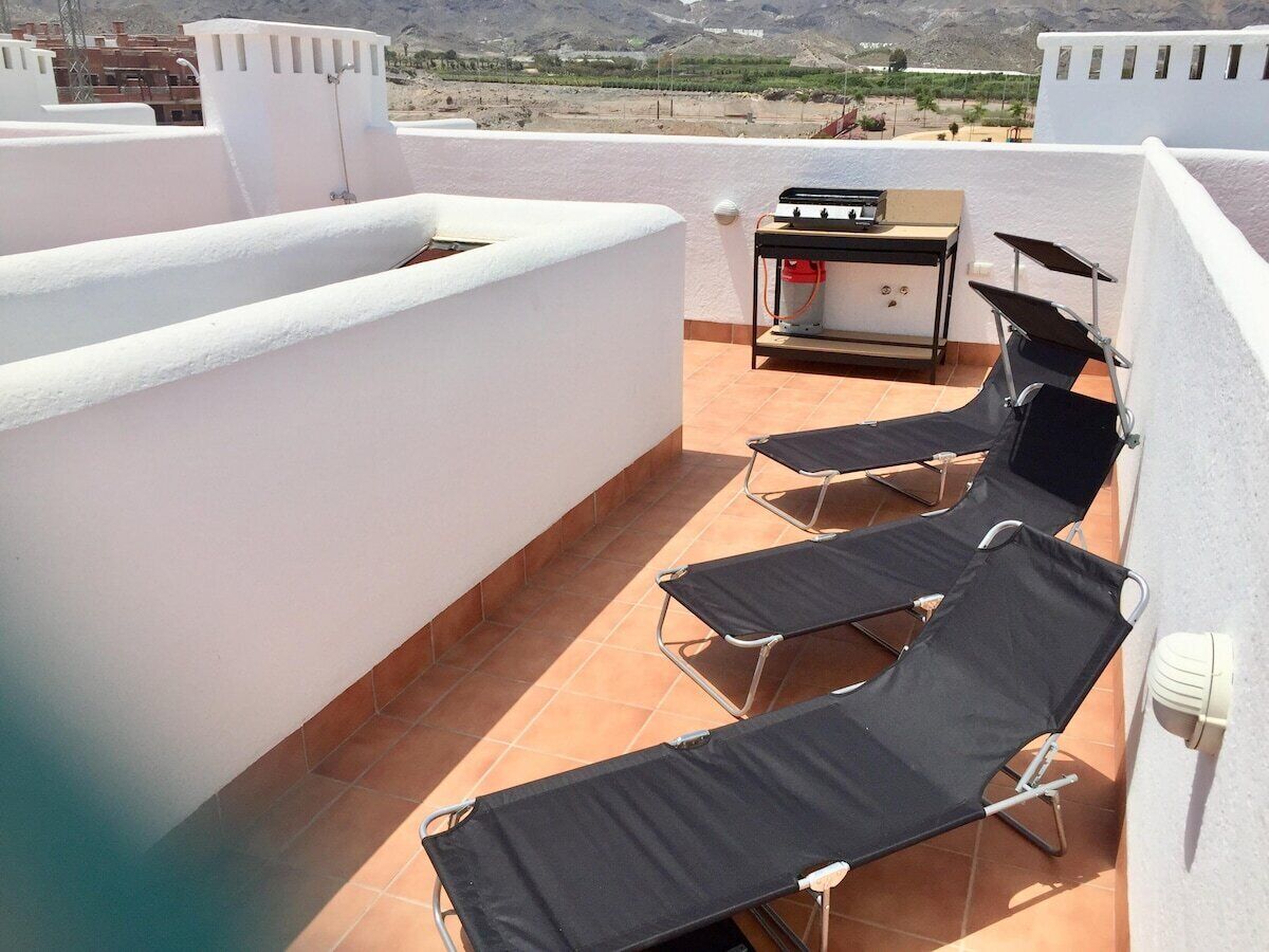 Eine Dachterrasse mit schwarzen Liegestühlen, einem Grill und Blick auf die umliegende Landschaft.
