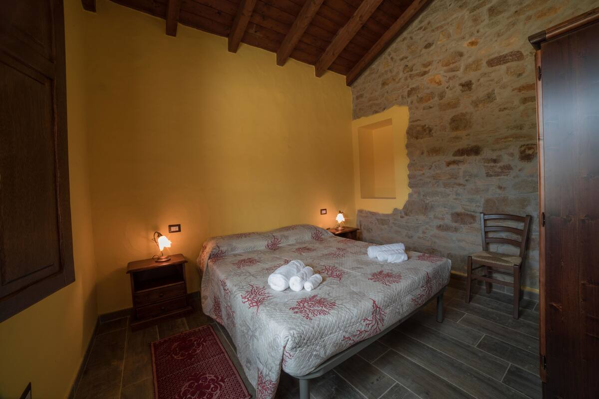 A bedroom with a stone wall, a bed with a floral bedspread, two bedside lamps, a wooden chair, and a red rug.