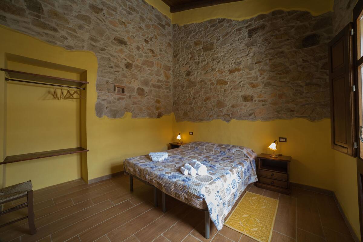 A bedroom with a stone wall, a bed with a patterned bedspread, a bedside table with a lamp, and a bench.