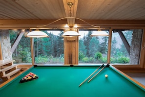 Une table de billard recouverte d’un drap vert, entourée de bois, et offrant une vue sur une forêt grâce à de grandes fenêtres.