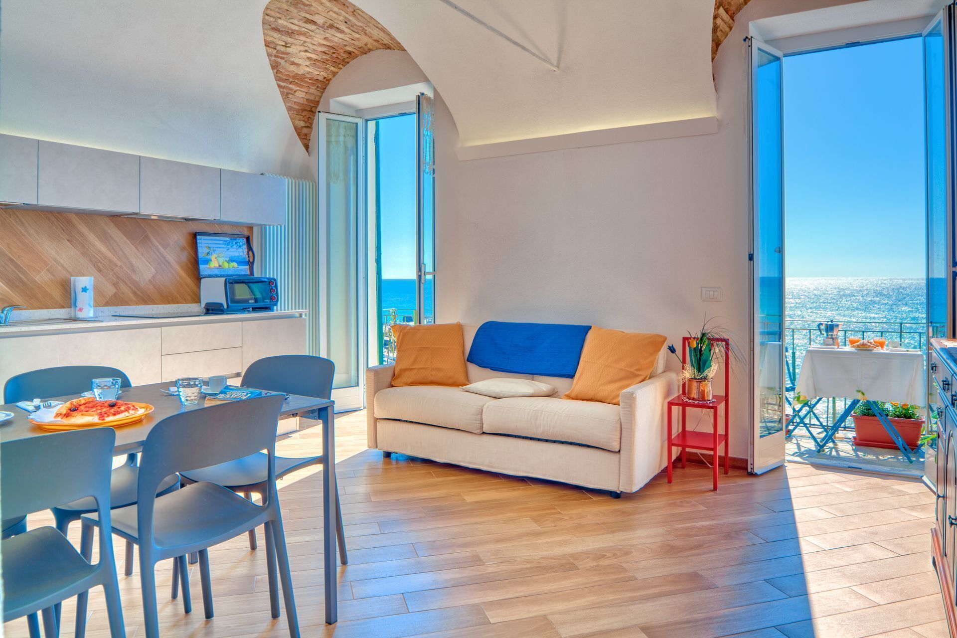Una sala moderna con sofá, mesa de comedor y sillas, una zona de cocina con microondas, y un balcón con mesa y sillas.