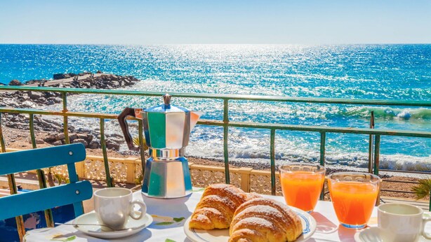 Een tafel gedekt met croissants, koffie en sinaasappelsap, met uitzicht op een rotsachtige kustlijn en de zee.