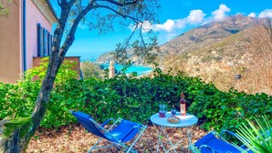 Un patio con due sedie blu, un tavolo con una bottiglia, un boccale e un piatto di cibo, con vista su una cittadina costiera e montagne.