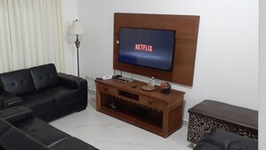 Ein Wohnzimmer mit einem schwarzen Ledersofa, einer Holzunterhaltungsecke mit Fernseher und 'NETFLIX'-Anzeige, einer Lampe und einem kleinen Tisch mit einer Figur.