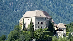A historic stone building with multiple windows and a sloped roof, surrounded by lush greenery and forested hills. - Mittendorf Klimaresidence (Schenna)