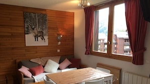 A cozy room with a wooden wall, a sofa with cushions, a wooden table, and a window with curtains. - Studio, at the foot of the Belle Plagne slopes, sleeps 4, wifi included. (La Plagne Tarentaise)