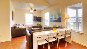 A living room with a ceiling fan, a TV, a painting, a dining table with chairs, and a window with blinds.