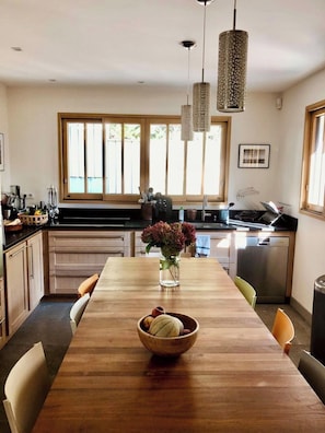 A modern kitchen with a wooden dining table, hanging pendant lights, and a view of the outdoors through large windows.