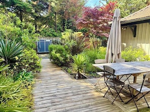 A wooden deck with a table and chairs, surrounded by lush greenery and a building in the background.