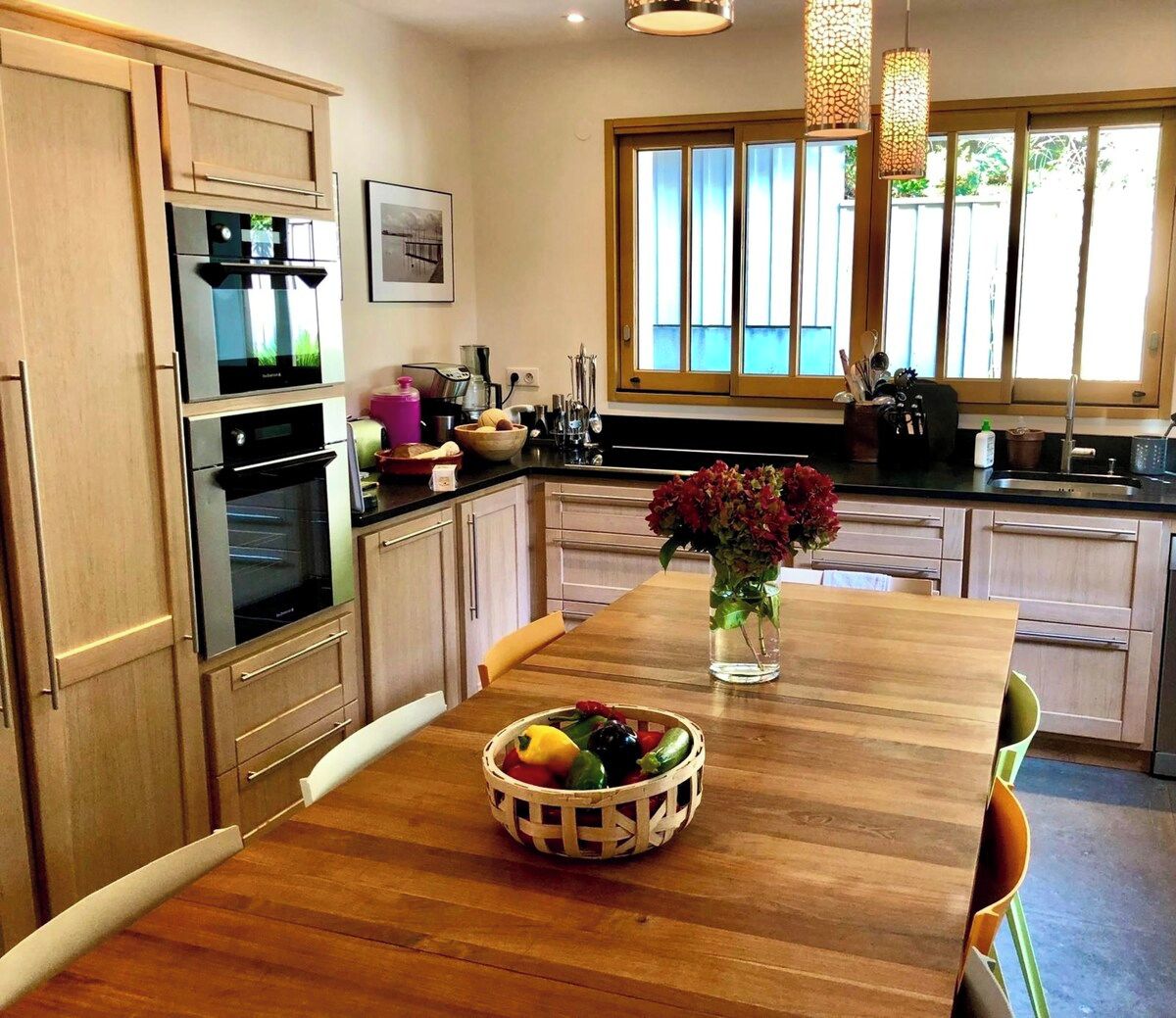 A modern kitchen with wooden cabinets, stainless steel appliances, and a wooden dining table with a fruit bowl and vase.