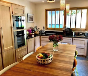 A modern kitchen with wooden cabinets, stainless steel appliances, and a wooden dining table with a fruit bowl and vase.