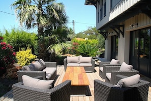 An outdoor patio with wicker furniture and a wooden table, surrounded by lush greenery and a building with large windows.