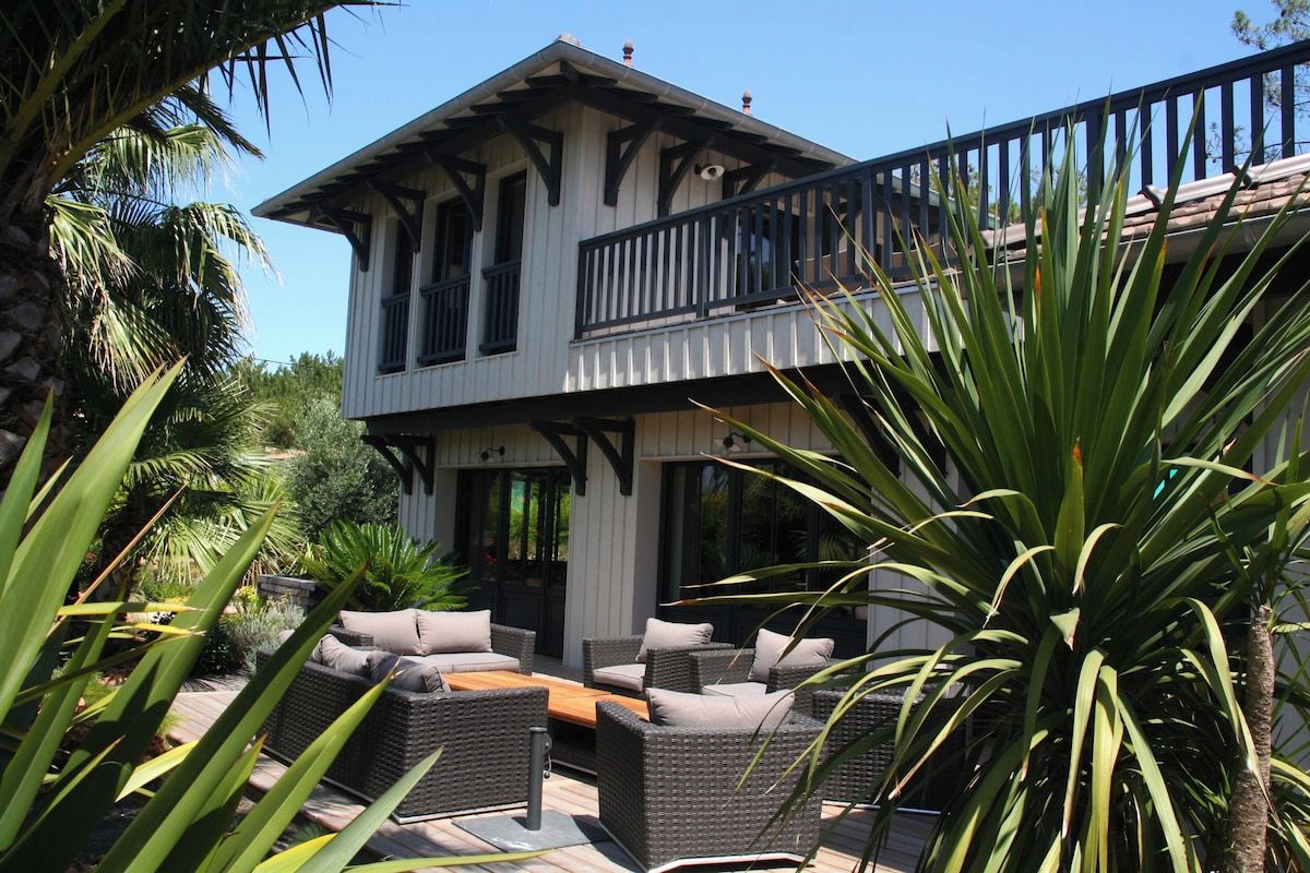 A two-story building with a balcony, outdoor seating area with wicker furniture, and tropical plants.