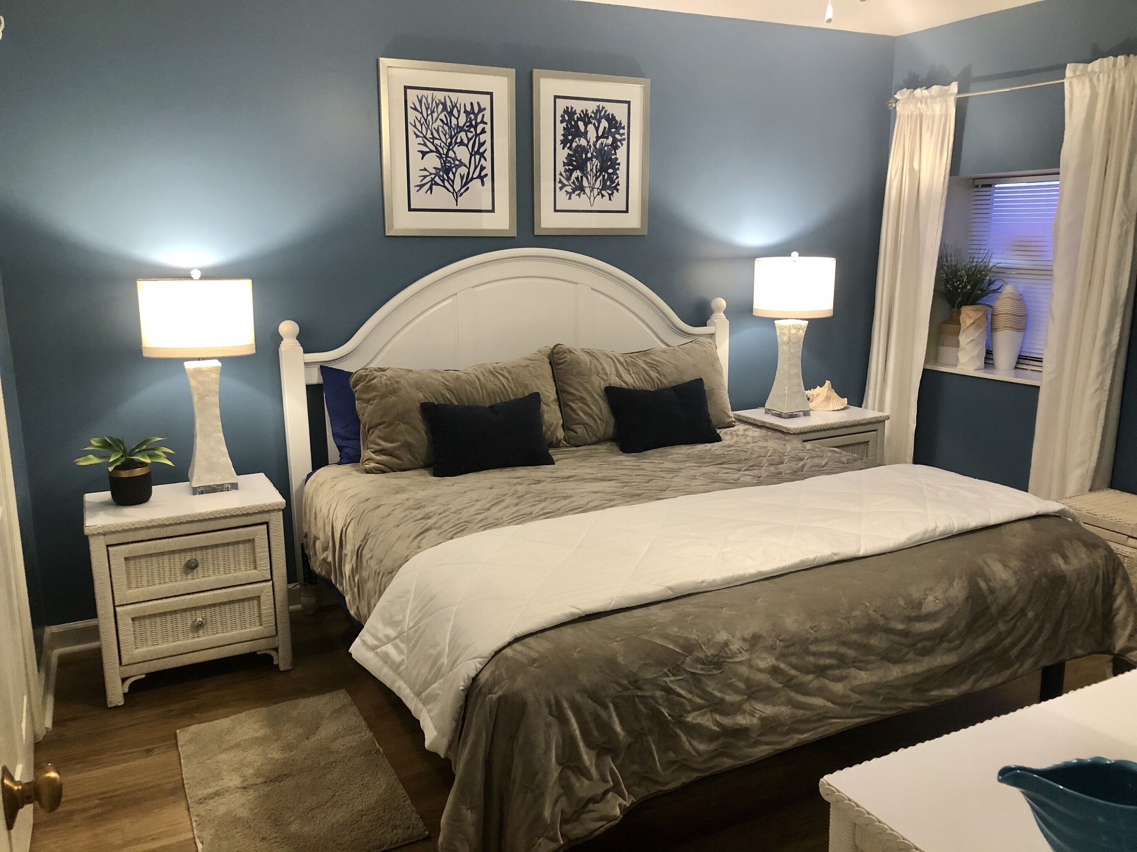 Une chambre à coucher comprenant un lit, deux tables de chevet avec des lampes, une commode, une fenêtre avec des rideaux et deux œuvres d’art murales.