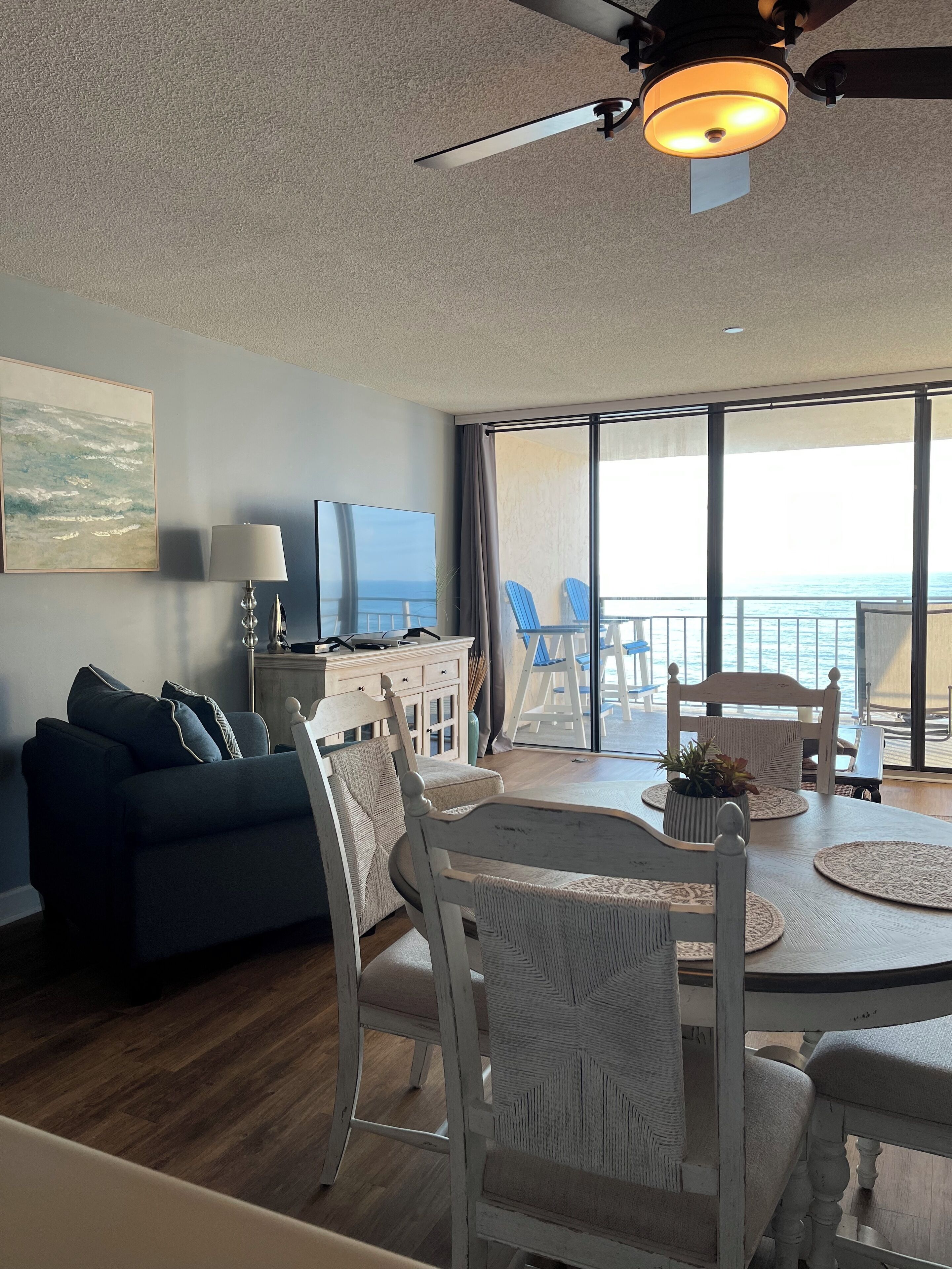 A modern living room with a sofa, dining table, chairs, and a TV. A balcony with a view of the sea is visible through large windows.
