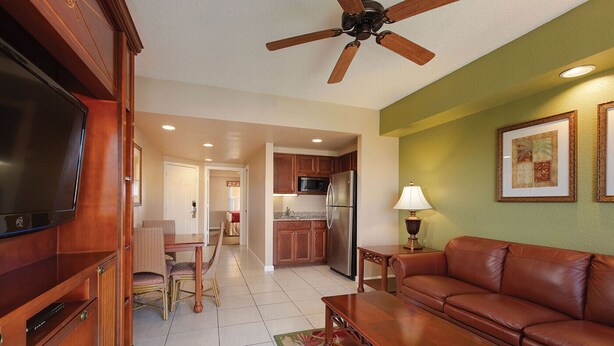 A living room with a brown leather sofa, wooden coffee table, and a TV on a wooden cabinet. There is a ceiling fan, a kitchen with a refrigerator, and a dining area with a table and chairs. - 7 Days / 6 Nights Gateaway (Kissimmee)