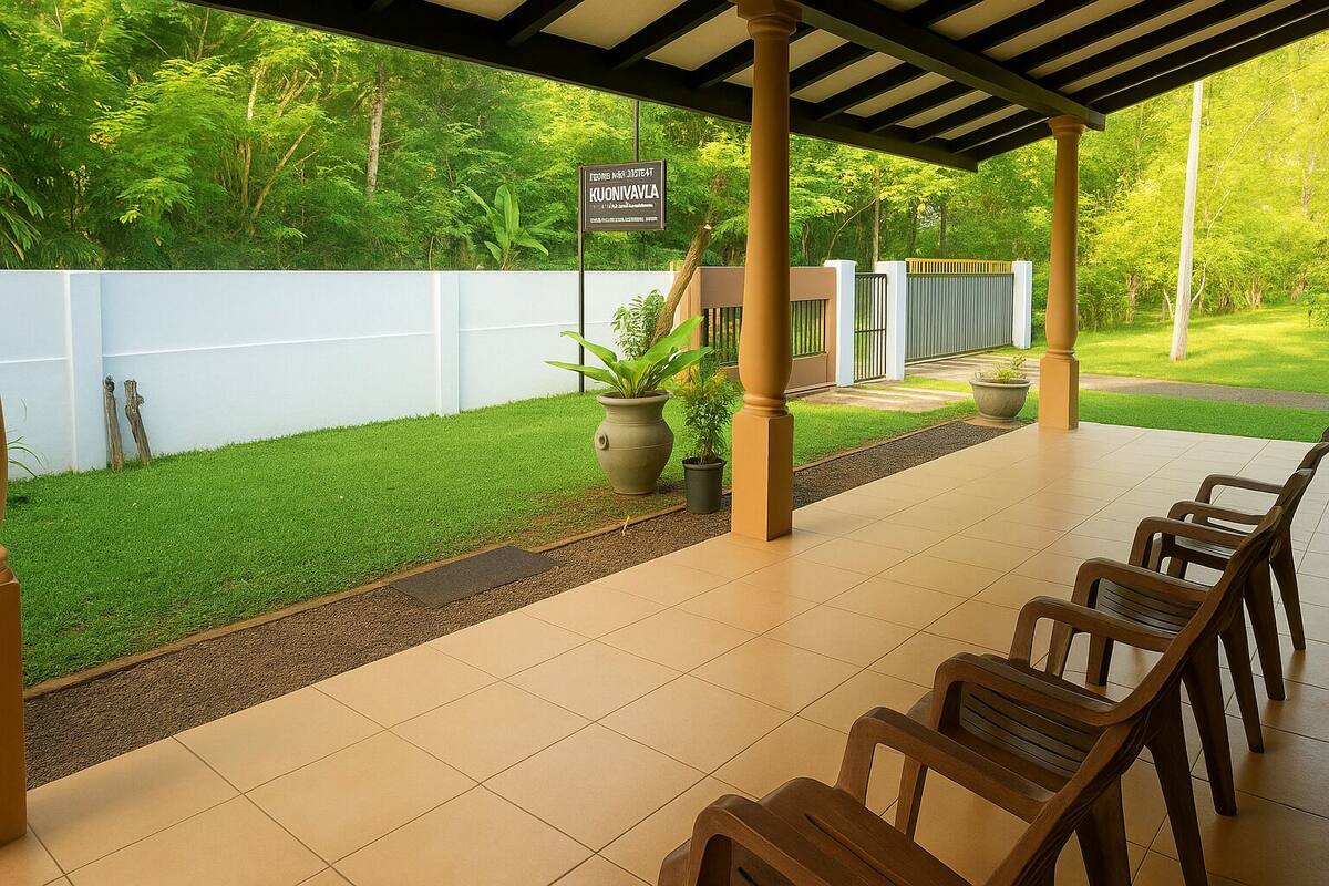 A covered outdoor seating area with a tiled floor, wooden chairs, potted plants, and a white wall with a sign.